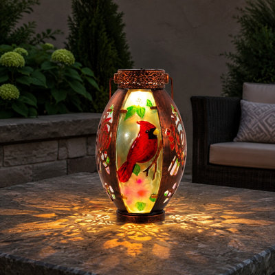 A waterproof Cardinal solar tabletop lantern combines Metal & Stained Glass with a Hollowed-Out Design for decorative use in yards, patios, gardens, and pathways. For outdoor use.