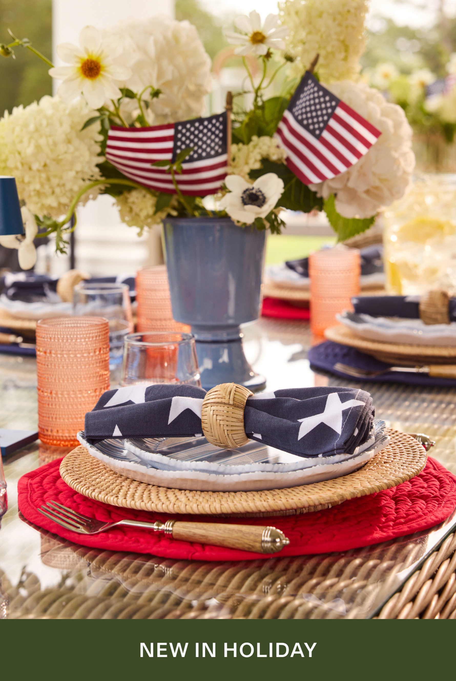 A patriotic 4th of July table setting featuring American flags in a blue vase with white flowers, a star-patterned navy napkin with wicker ring, red and natural woven placemats, and coral glassware. Text reads 'NEW IN HOLIDAY'.