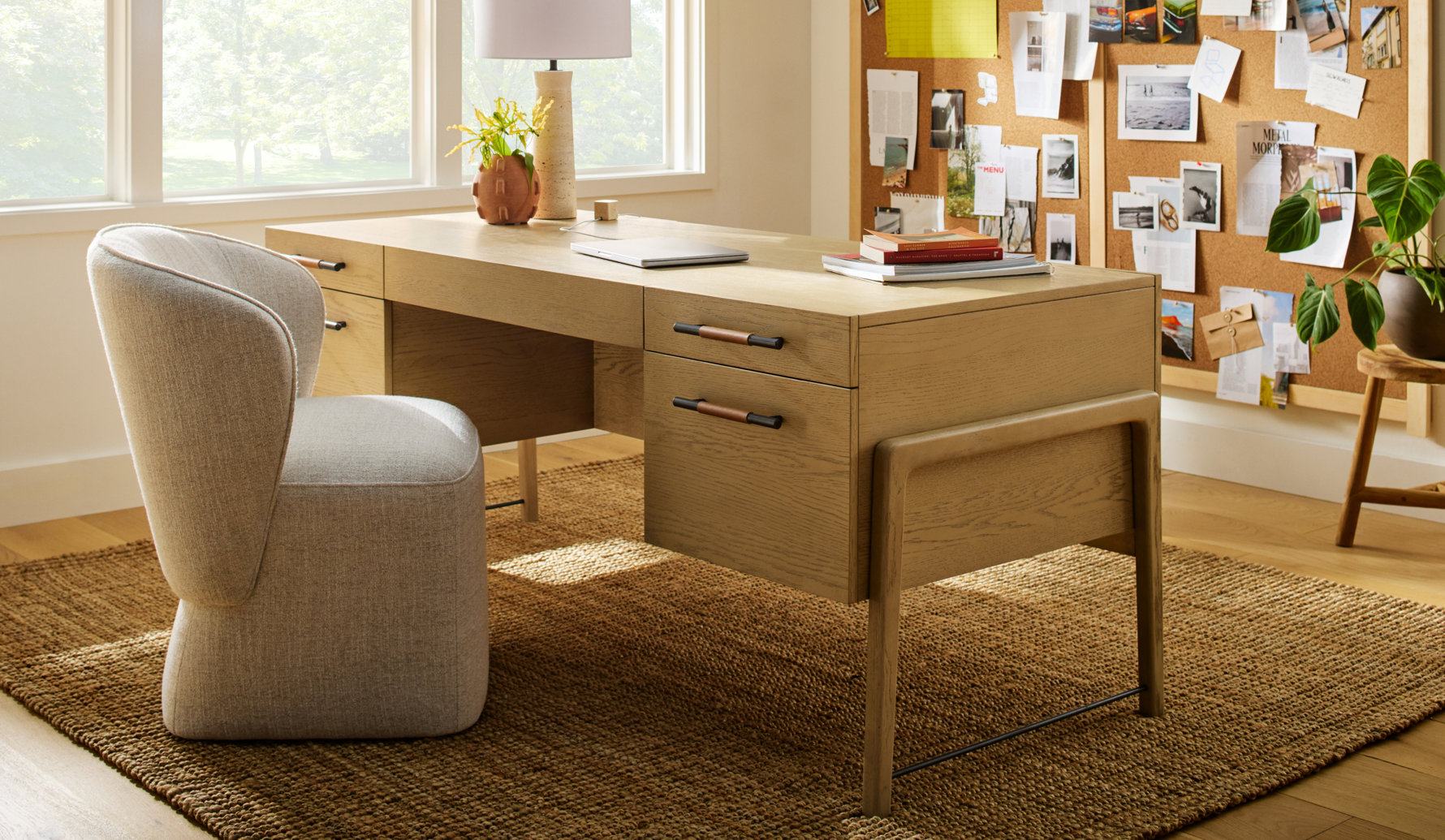 A light oak executive desk with black and leather drawer pulls sits on a natural fiber rug next to a modern upholstered office chair, with a cork inspiration board on the wall.