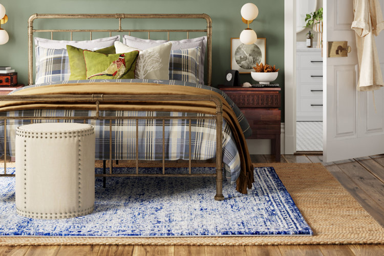 cozy bedroom with a jute rug beneath a blue area rug, antiqued brass bed, plaid bedding, and a white nailhead-trim pouf