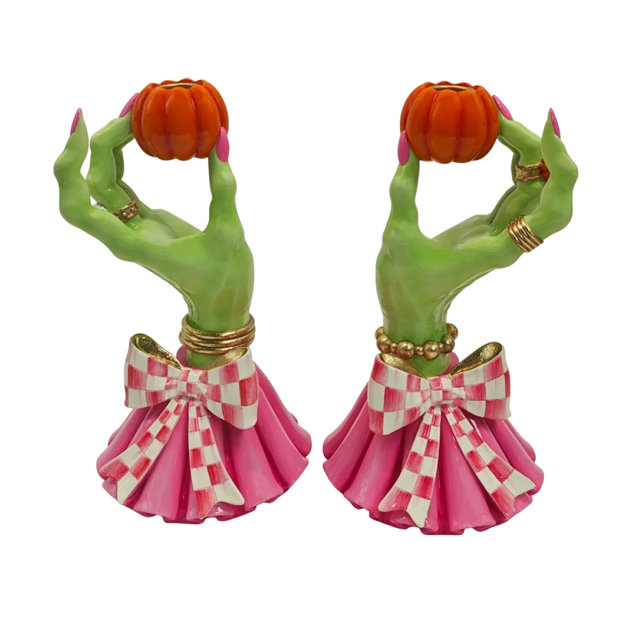 Two decorative candle holders shaped like green witch hands wearing gold rings and pink dresses with white checkered bows, each holding a small orange pumpkin.