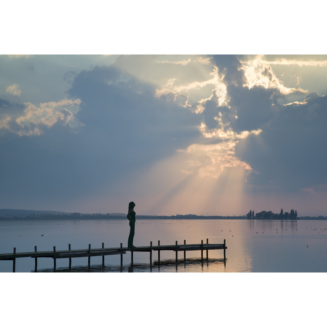 Silhouette einer jungen Frau, die auf einem Steg am Seeufer in einem Sonnenstrahl steht