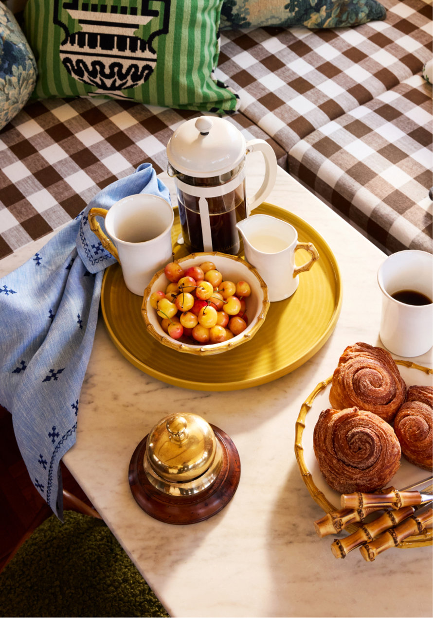 Coffee and breakfast pastries served on a yellow tray with a bowl of cherries, a French press, and a brass bell on a marble tabletop with gingham seating.