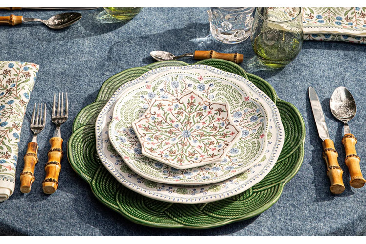 An elegant, garden-inspired table place setting arranged on a blue textured tablecloth. The stack of patterned porcelain dinnerware features a scalloped green woven-look charger plate at the base, topped with two tiers of white plates decorated with delicate blue and green botanical prints, and finished with a small star-shaped floral dish in the center. The setting is flanked by forks, a knife, and spoons with natural bamboo handles, alongside a floral patterned cloth napkin and green glassware.