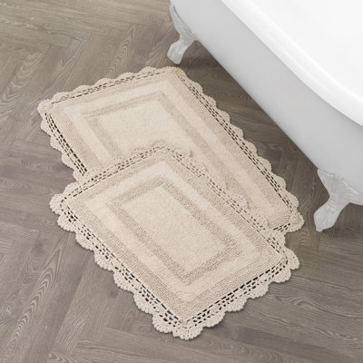 Two beige cotton bath rugs with scalloped crochet borders arranged on a wood floor.