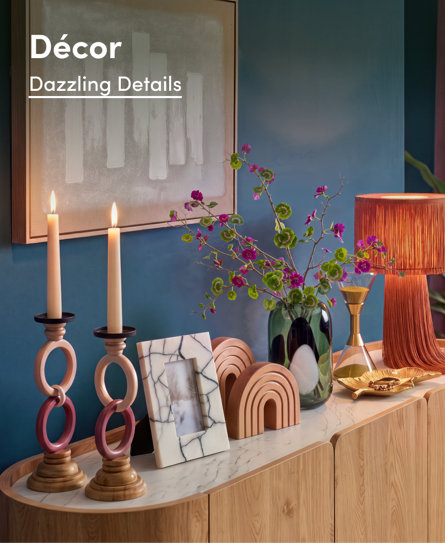 Décor: Dazzling Details featuring a console table styled with modern candles, a marble-effect frame, sculptural arches, a glass vase with purple flowers, and a fringed lamp against a deep blue wall.