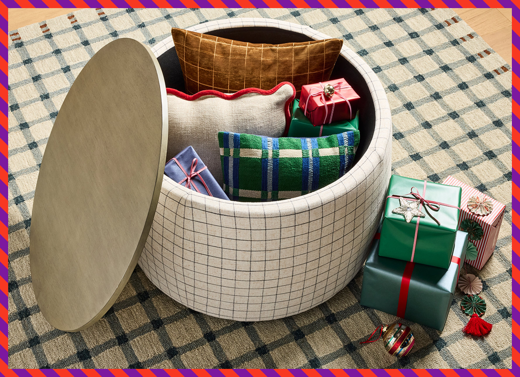 A round cream-colored storage ottoman with a black grid pattern, shown with its lid open to reveal decorative pillows and wrapped holiday gifts inside, resting on a matching plaid area rug with additional wrapped gifts nearby.