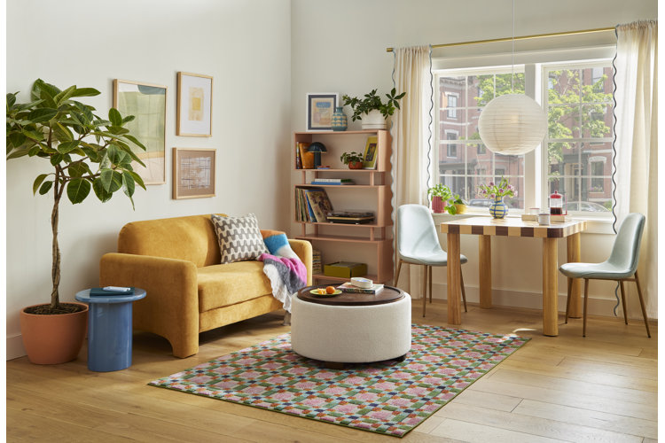 A vibrant, modern living and dining space featuring a yellow loveseat, a colorful checkered rug, a cream storage ottoman coffee table, a wooden dining table by a large window with two light blue chairs, and a potted rubber tree plant