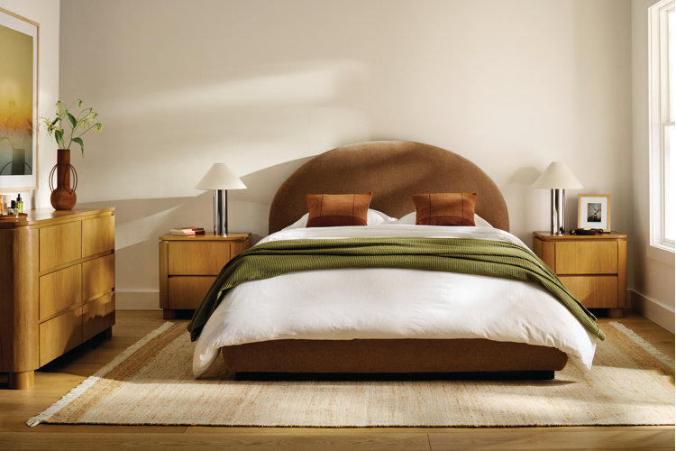 A serene and modern bedroom featuring a brown upholstered bed with a large, arched headboard. The bed is made with crisp white linens, a folded olive green textured blanket across the middle, and two burnt orange accent pillows.