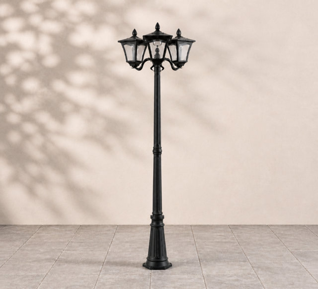 Traditional three-head black outdoor lamp post with glass panels standing on a tiled patio.