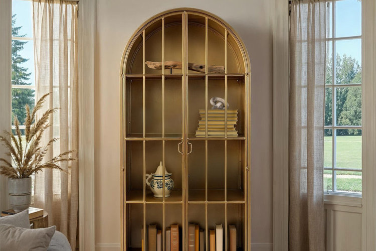 Arched gold metal curio cabinet with glass doors displaying books and decorative accents in a bright living room