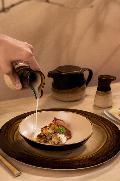 A hand pours cream from a rustic, two-toned ceramic pitcher into a bowl of food set on a matching dinner plate, accompanied by a creamer and a teapot on a wooden table.