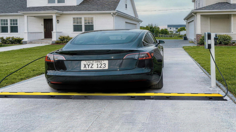 Véhicule électrique noir stationné dans une entrée résidentielle avec un passe-câble jaune et noir recouvrant un câble de recharge.