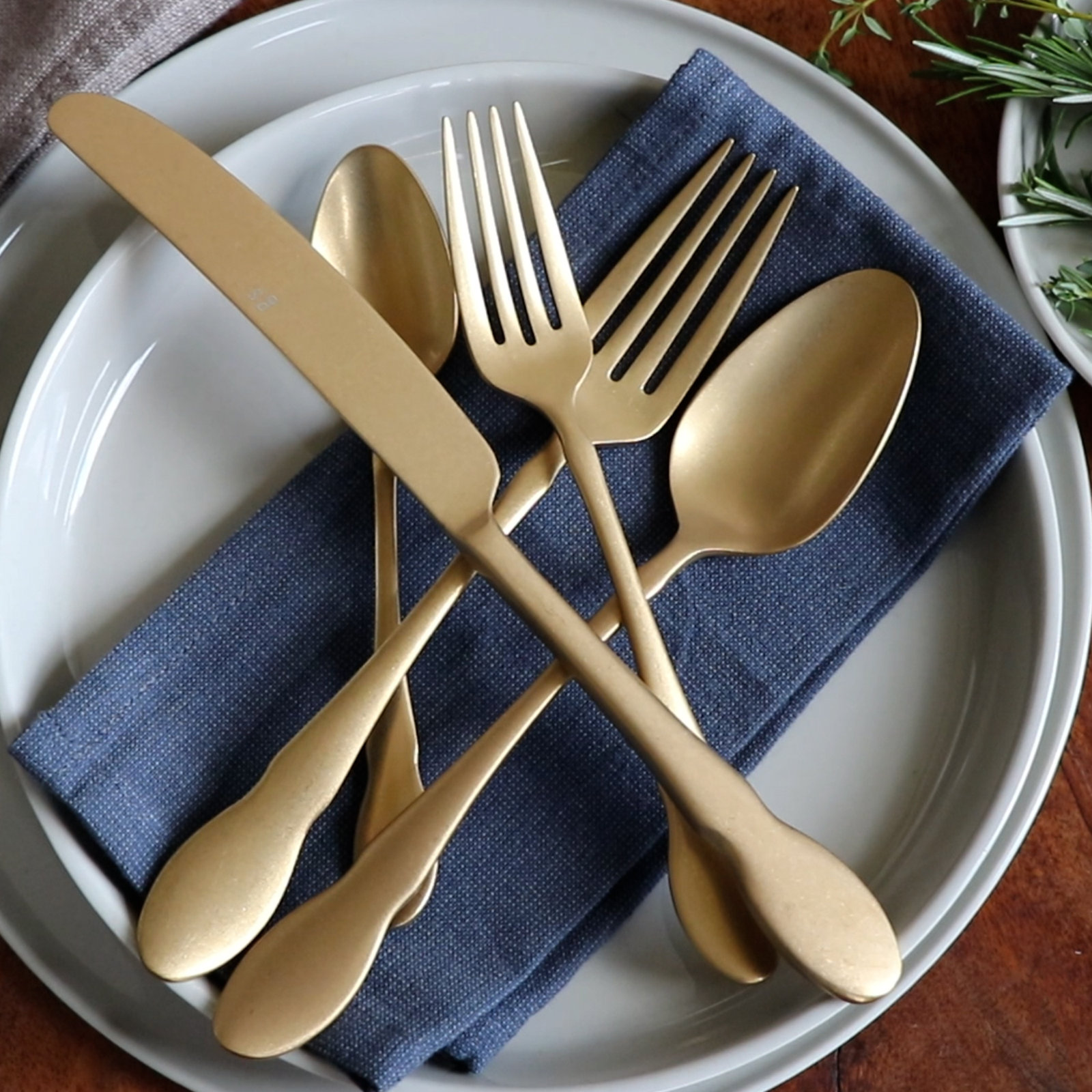 Gold-toned stainless steel flatware set with a matte finish, displayed on a blue fabric napkin and gray ceramic plates.
