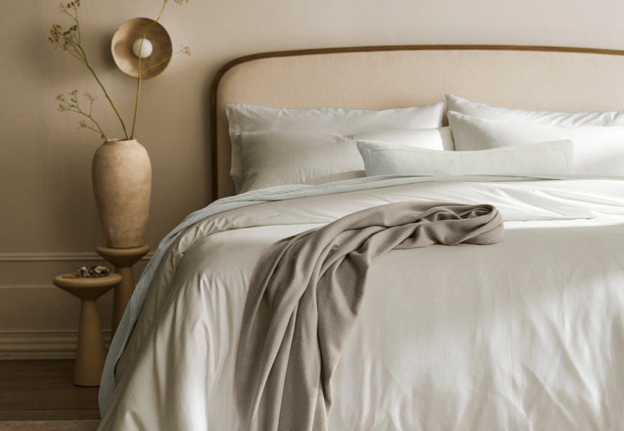 A neutral-toned bedroom scene featuring a bed with white bedding, layered with a light blue quilt and a textured beige throw blanket, next to a minimalist bedside table and a vase with sprigs.