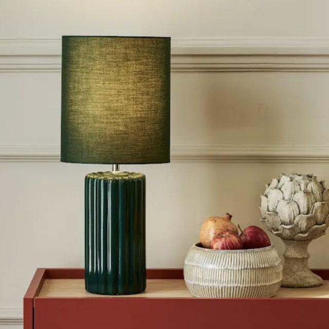 Dark green fluted ceramic table lamp with a matching fabric drum shade sitting on a red sideboard next to a decorative bowl of fruit and an artichoke ornament.