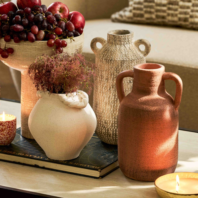 A collection of textured decorative vases and a fruit bowl filled with grapes and apples sit on a living room coffee table alongside lit candles and a book.