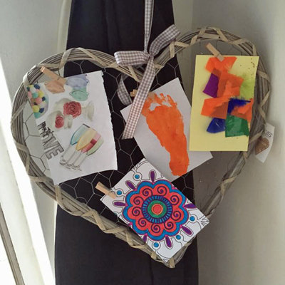 A heart-shaped frame with a wire backing displaying four pieces of children's artwork