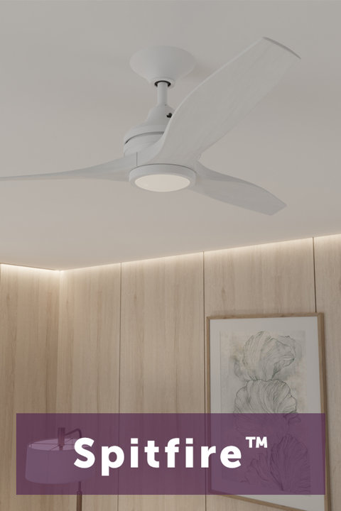 Modern white three-blade ceiling fan with an integrated light kit mounted on a white ceiling above a wall with wood paneling and framed art, featuring the text Spitfire TM.
