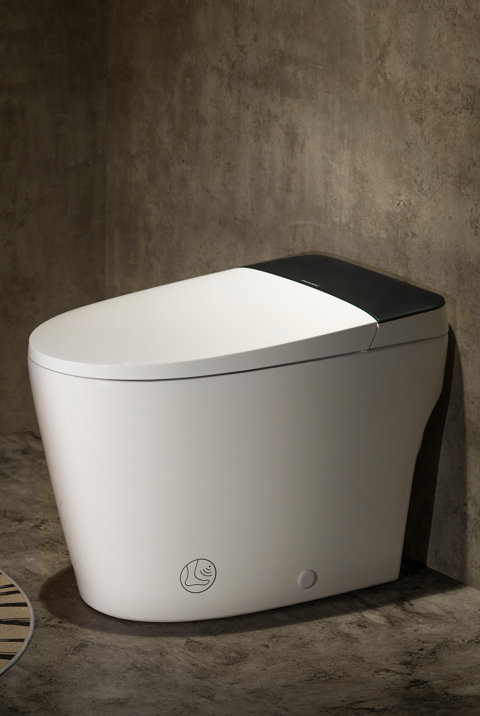 Modern white smart toilet with a black lid and foot sensor icon on a concrete floor against a concrete wall.