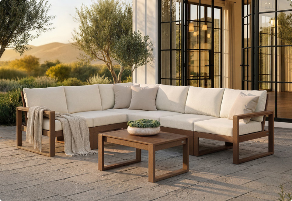 Outdoor sectional sofa with cream cushions and a matching low coffee table, made of brown POLYWOOD lumber, on a stone patio with a sunset landscape behind it