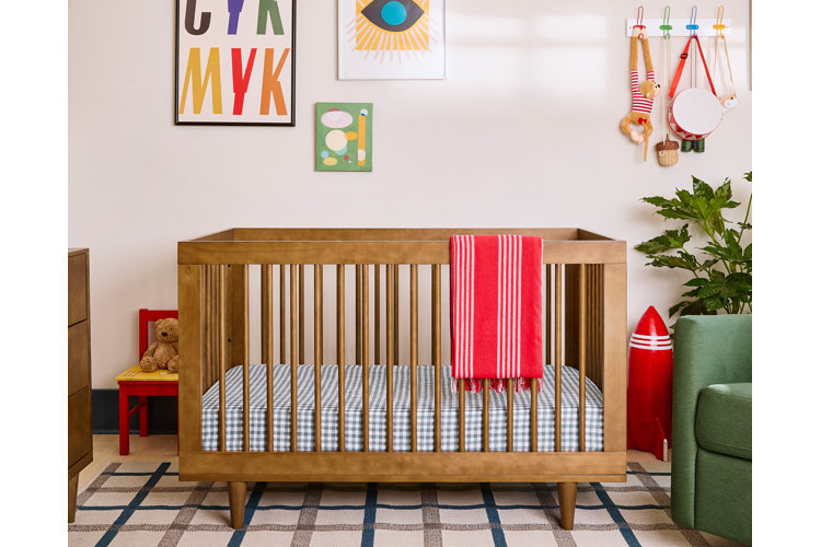 brown wood crib in a nursery. blue checkered crib sheet. whimsical wall art and a small wood chair with a teddy bear on it