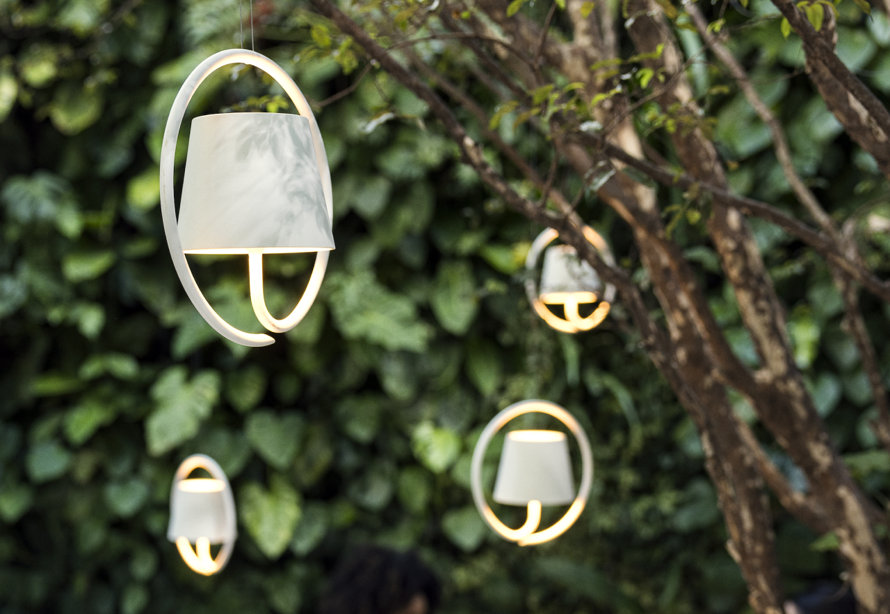 Three modern, white, circular pendant lights with tapered shades hanging outdoors in front of a lush green foliage wall.
