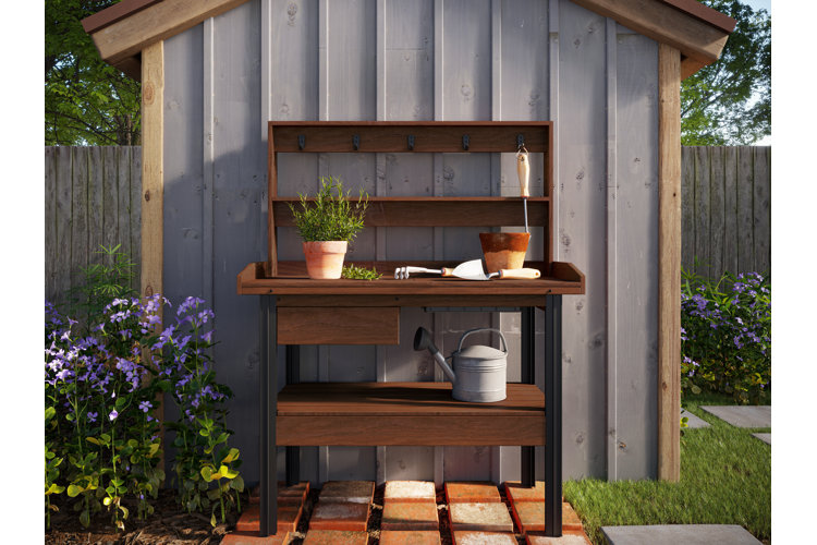 Wood potting bench up against a garden shed with a watering can and terracotta pots, one with a rosemary plant inside.