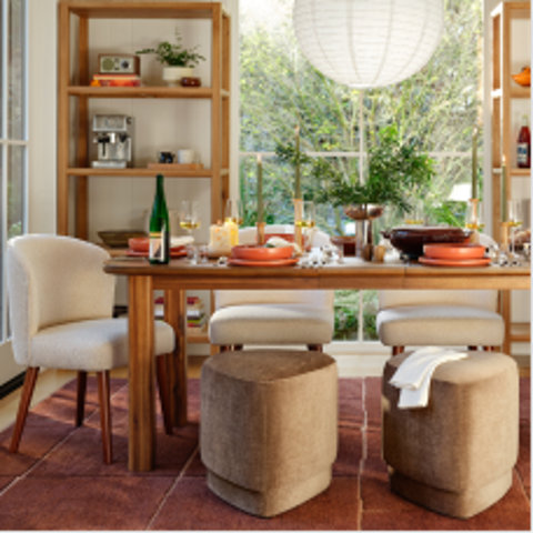 Dining room set featuring a wooden table with cream upholstered chairs and two beige triangular ottomans placed on a terracotta rug in front of a window.