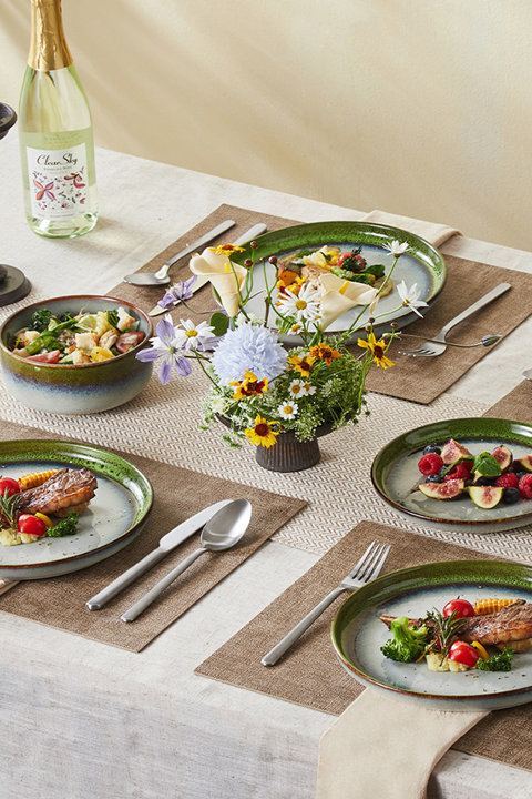 A beautifully set dining table featuring green and blue reactive glaze dinnerware, a centerpiece of wildflowers, and plates served with grilled meat, vegetables, and fresh fruit.