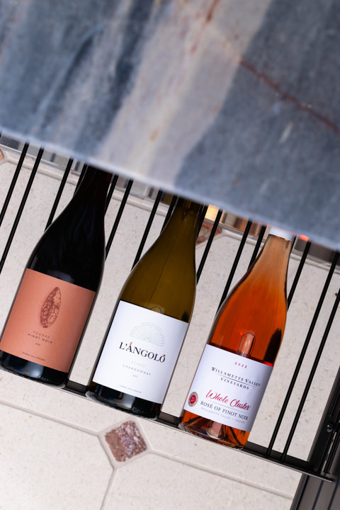 Three bottles of wine arranged on a dark metal rack, including Foudre Pinot Noir, L'Angolo Estate Chardonnay, and Willamette Valley Vineyards Whole Cluster Rosé of Pinot Noir.