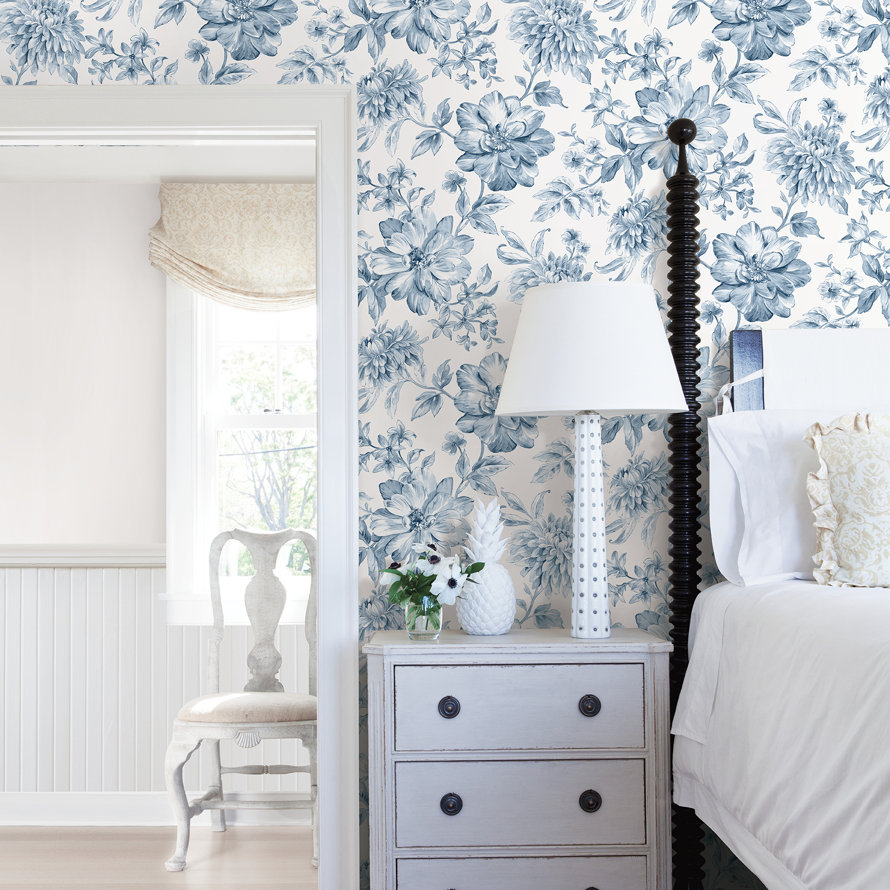 Bedroom featuring a floral blue and white wallpapered wall, a white bedside table with a lamp, and a view into an adjoining room with a wooden chair.