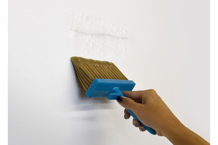 A person's hand holding a paint brush and applying wallpaper paste to a bare wall.