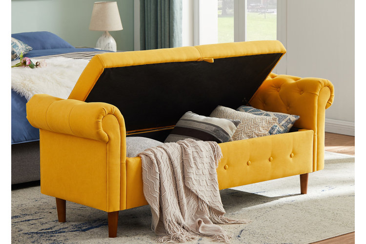 bright yellow tufted storage bench holding extra throw pillows and blankets in front of a blue bed