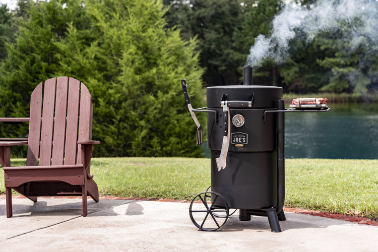 Vertical black smoker on a patio beside a lake.
