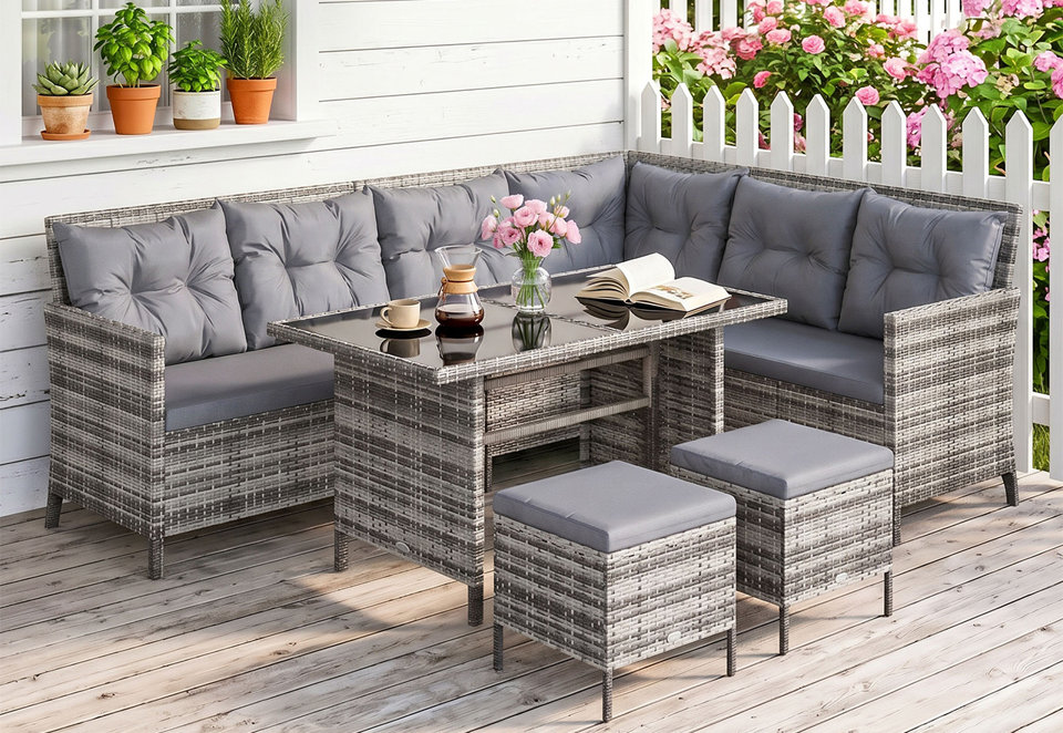 Grey rattan corner garden sofa set with a glass-topped dining table and two matching stools with grey cushions, set on a wooden patio against a white picket fence.