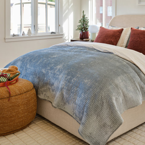 A modern bedroom featuring a cream-colored upholstered bed with green accent pillows, a burl wood nightstand topped with a small lamp, and a textured rust-colored rug.