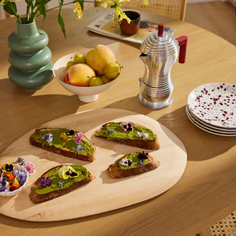 Avocado toast with edible flowers, fruit, and coffee maker on table.