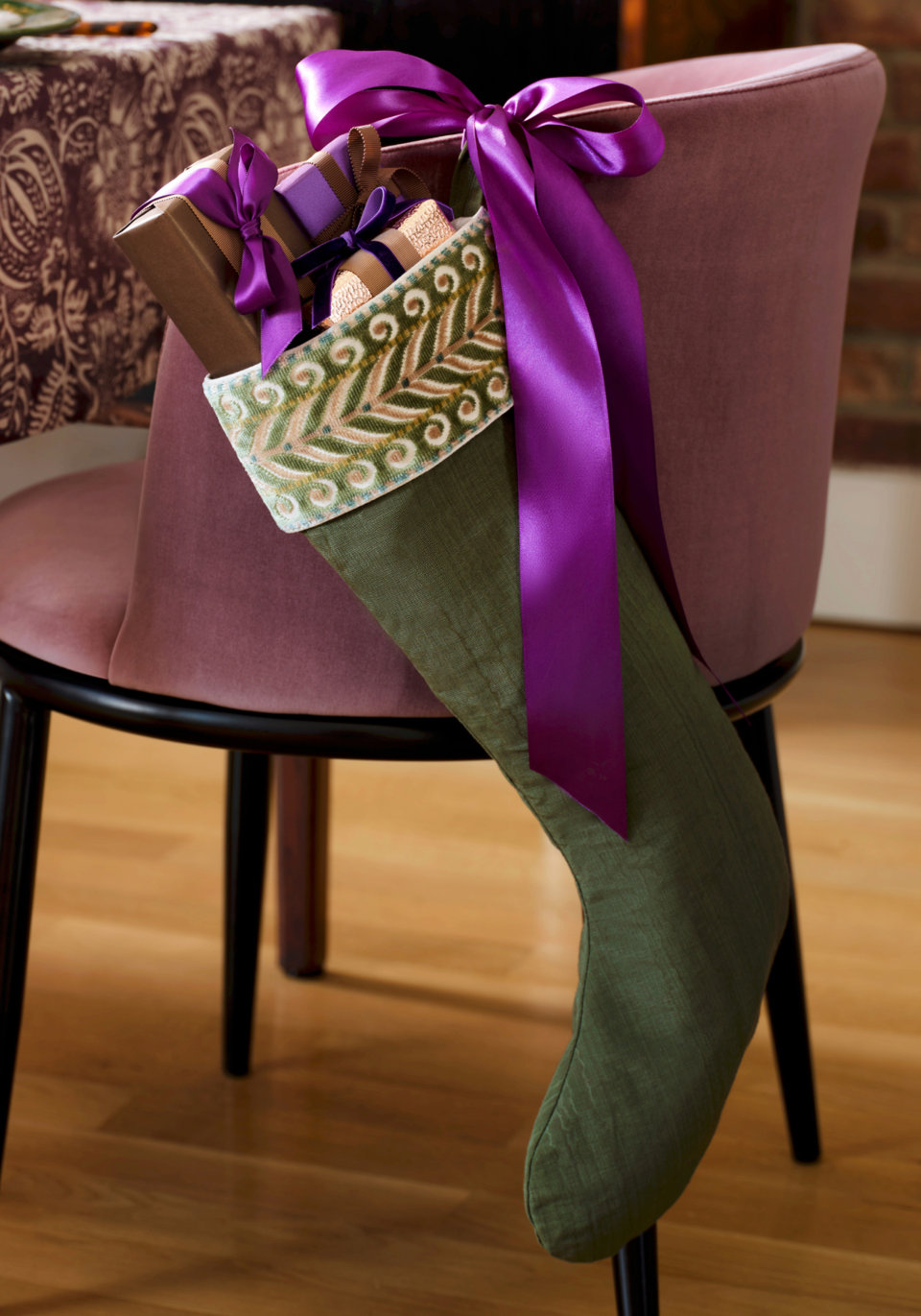 Green linen holiday stocking with a decorative embroidered cuff and a large purple satin bow hanging from a mauve velvet chair.