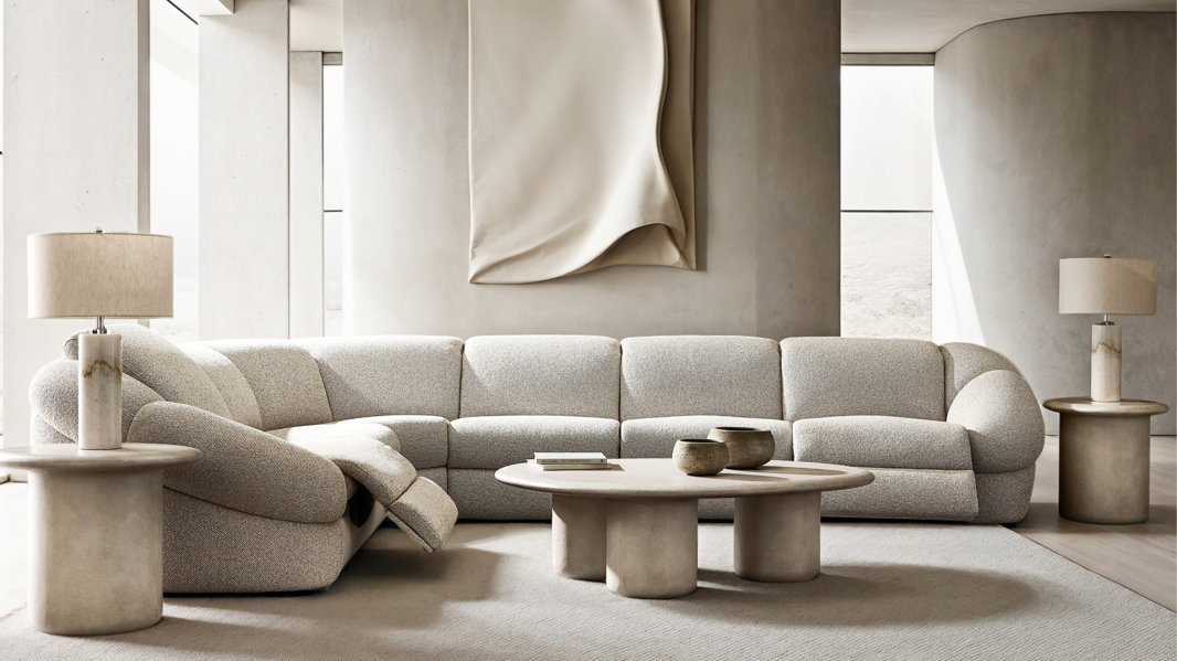 A modern living room featuring a curved, textured beige sectional sofa with a reclining seat, a low round stone coffee table with decorative bowls, and matching side tables topped with cylindrical lamps against a minimalist, neutral-toned wall with oversized fabric art.