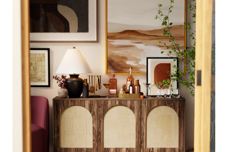 Close up of a wood and rattan sideboard being used as a home bar placed against a wall with brown abstract wall art.