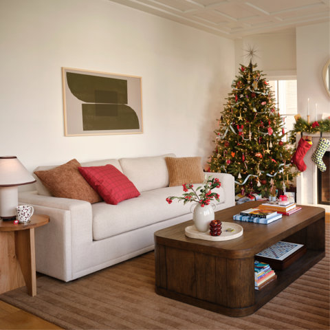 Cozy living room decorated for Christmas with a beige sofa, colorful throw pillows, a wooden coffee table, and a decorated Christmas tree near a fireplace.