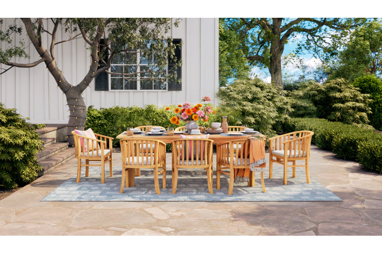 Wood outdoor dinner table heavily laden with dishes, surrounded by eight matching wood chairs. Blue outdoor area rug