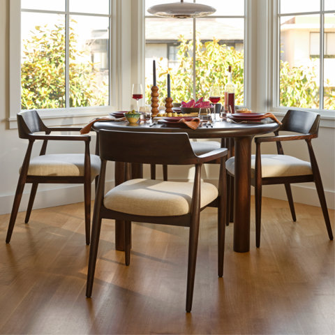 Round wooden dining table with three upholstered chairs in a sunlit breakfast nook.