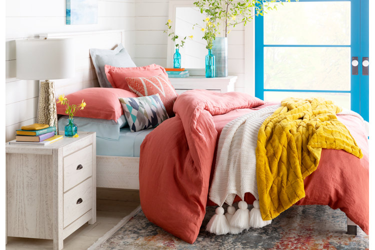 white bed frame with peach bedding set along with yellow and white throws next to a white nightstand