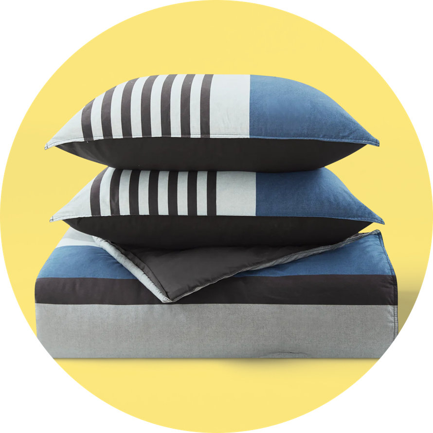 Blue and grey colorblock comforter and matching striped pillows stacked against a yellow circular background.