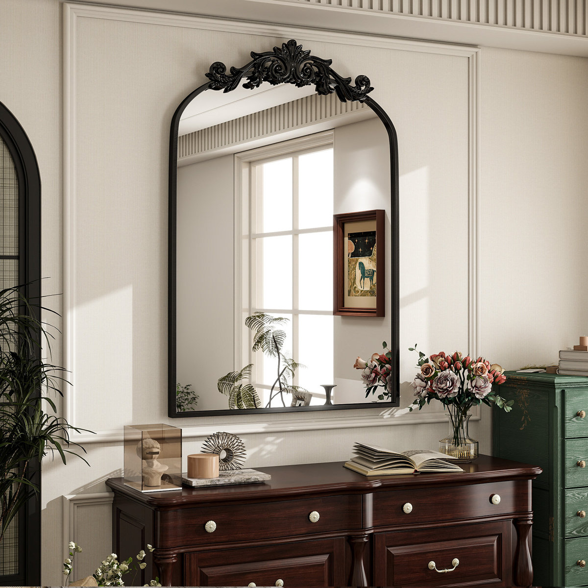 Large black-framed baroque mirror with ornate crest hanging above a dark wood dresser decorated with books, flowers, and minimalist decor in a softly lit room