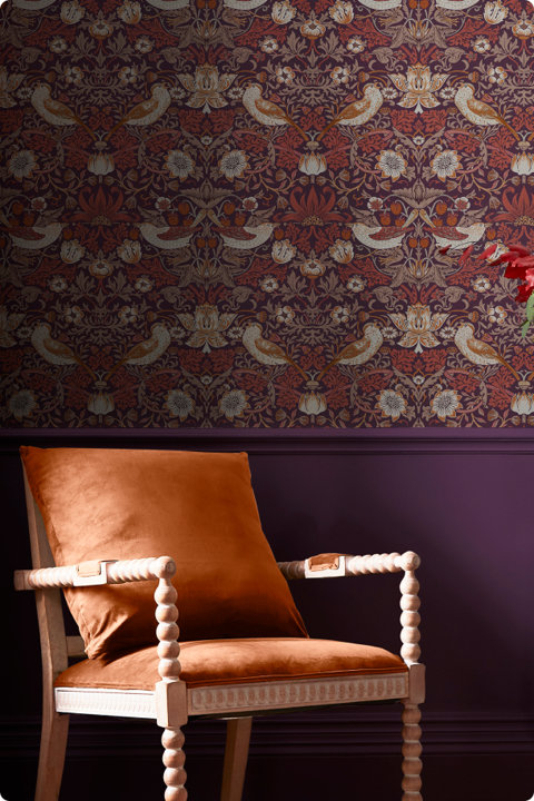 Rust orange velvet accent chair with a spindle frame positioned against a dark purple wall with ornate floral and bird patterned wallpaper.
