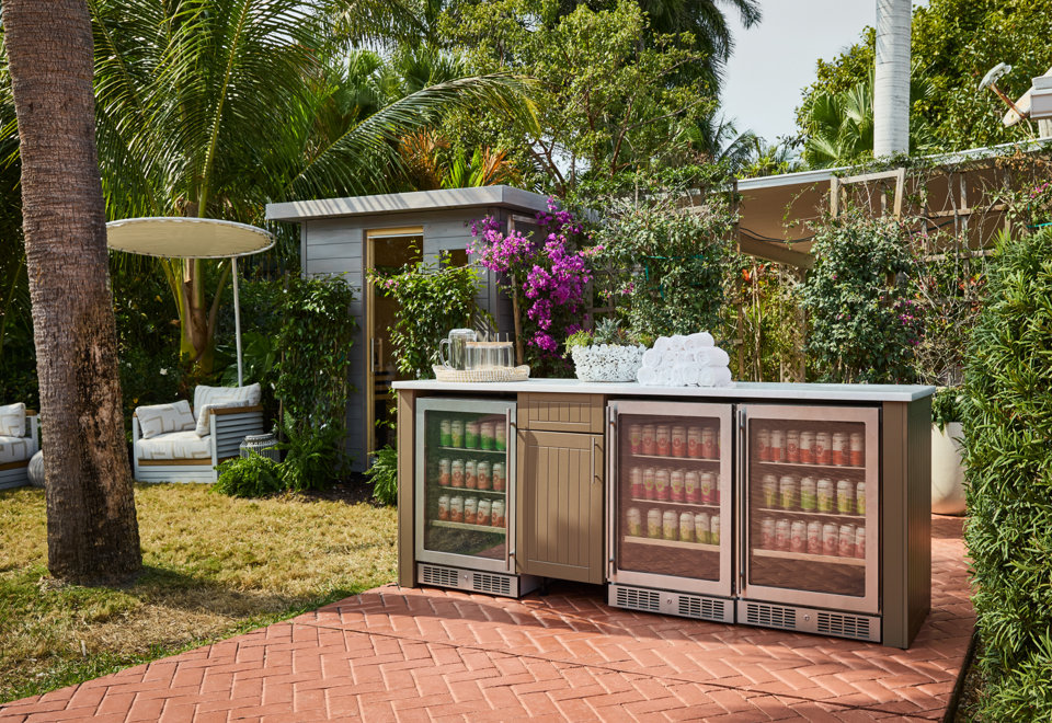 Outdoor kitchen island with a stone countertop, built-in stainless steel beverage coolers stocked with drinks, and a cabinet base set on a brick patio in a lush, tropical backyard with palm trees and a small sauna structure.