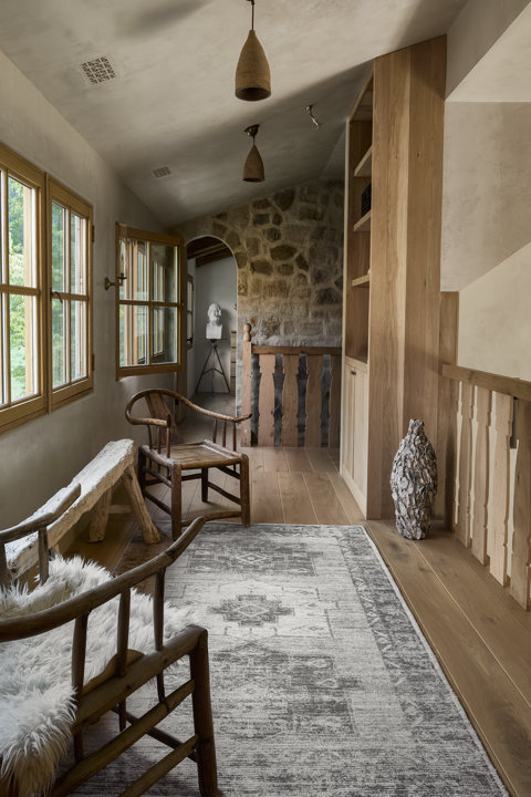 Couloir rustique avec murs en pierre, rampes en bois, un tapis gris et des chaises en bois ornées d'une jetée en fausse fourrure.